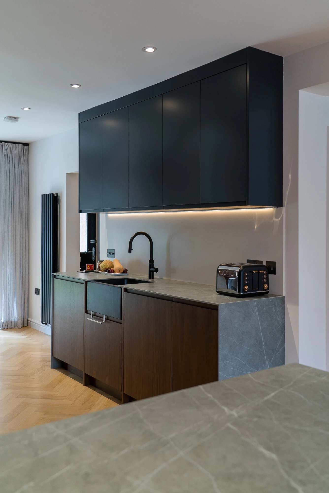 A modern kitchen with dark cabinets and a gray countertop. Under-cabinet lighting illuminates the area. The counter features a black faucet, a bowl of fruit, and an appliance. A curtain and radiator are visible in the background. The floor is light wood