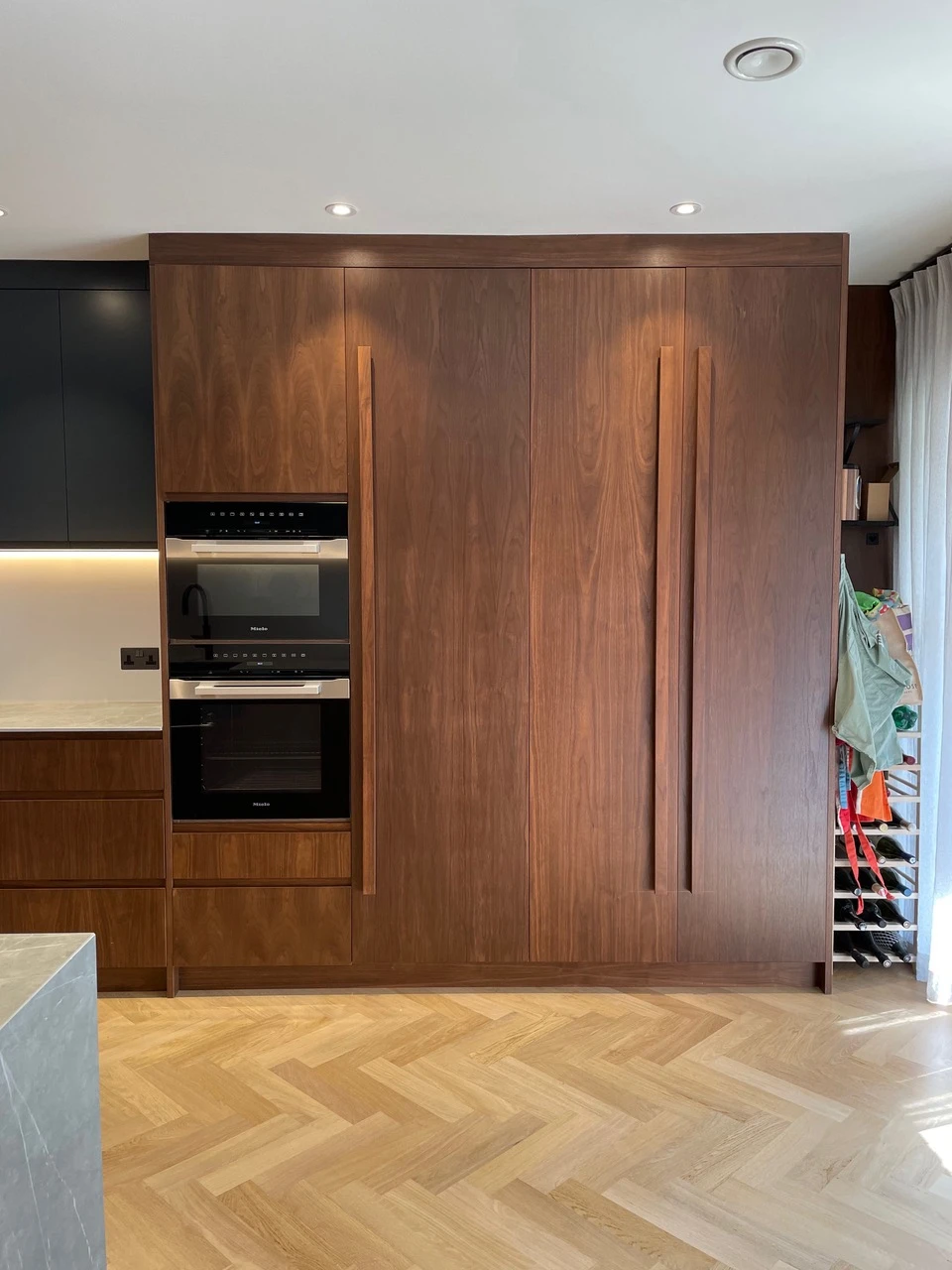 A modern kitchen with wooden cabinetry featuring double integrated ovens on the left. The cabinets have sleek, vertical handles. A countertop with an under-cabinet light is partially visible on the left, and an apron and bags hang from hooks on the right.