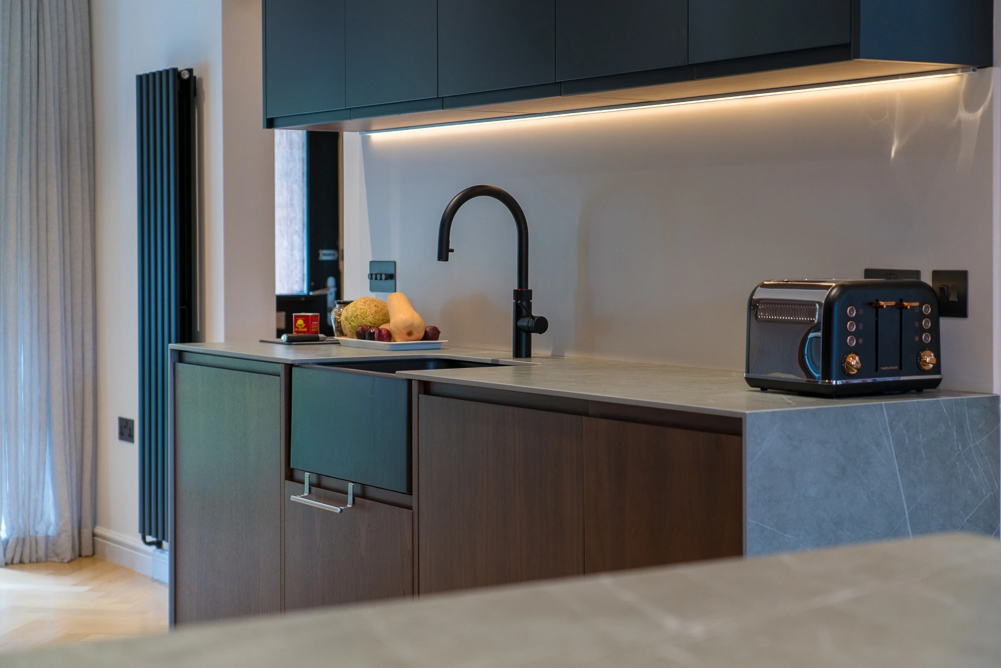 Modern kitchen with dark cabinets, a sleek black faucet, and under-cabinet lighting. A fruit bowl sits near the sink; a toaster is placed on the countertop to the right. Light grey countertops complement the dark wood cabinetry