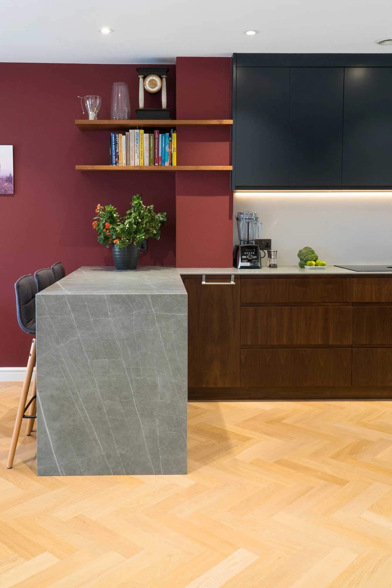 Modern kitchen with maroon walls, black upper cabinets, wooden lower cabinets, and light wood herringbone flooring. Gray marble countertop island with two high chairs, shelves with books, glassware, plants, a wall clock, and kitchen essentials on counter.