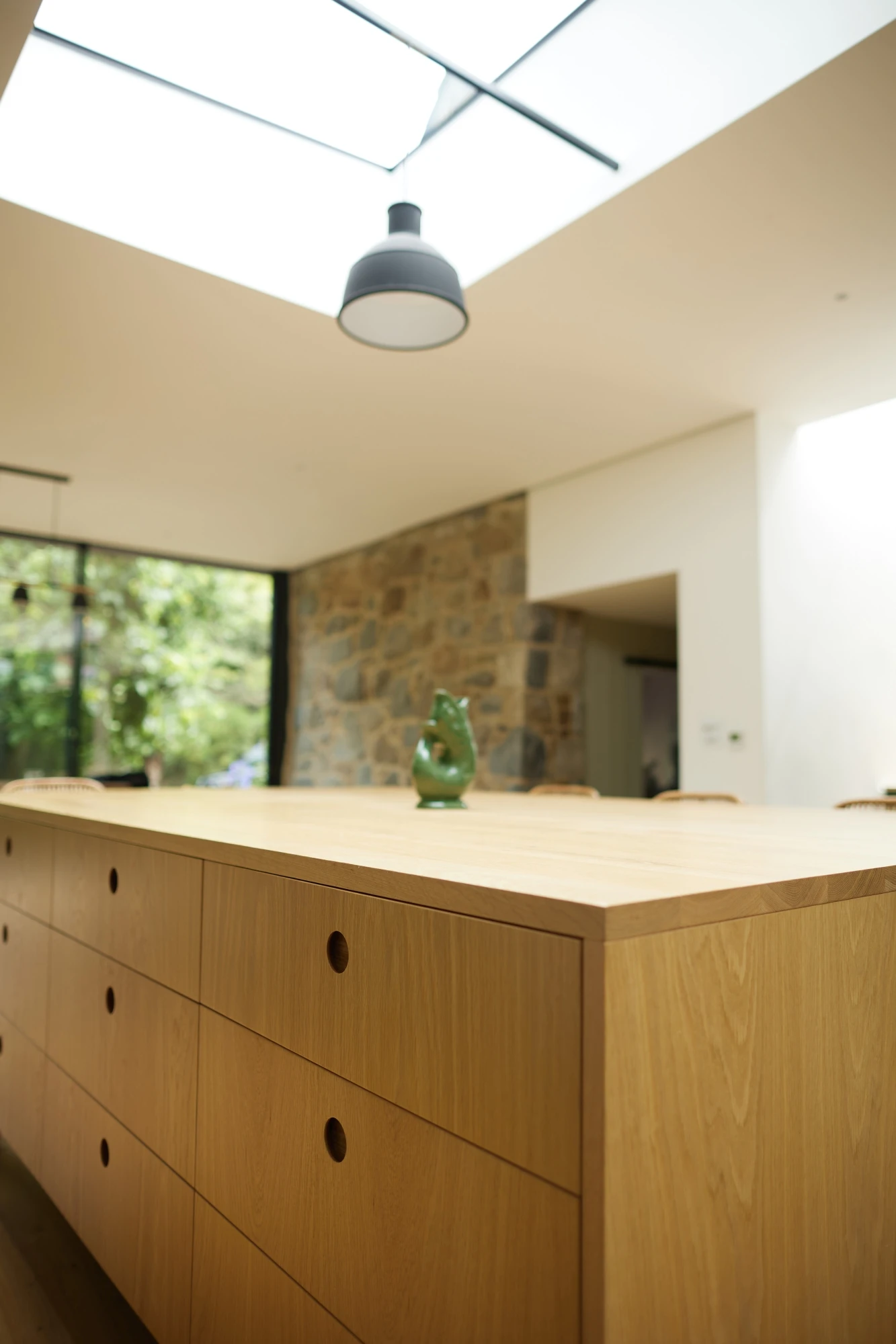 A modern kitchen with a wooden island featuring multiple drawers. A green decorative object sits on top of the island. The kitchen has a stone accent wall, large windows allowing natural light, and a skylight with a pendant light suspended from it.