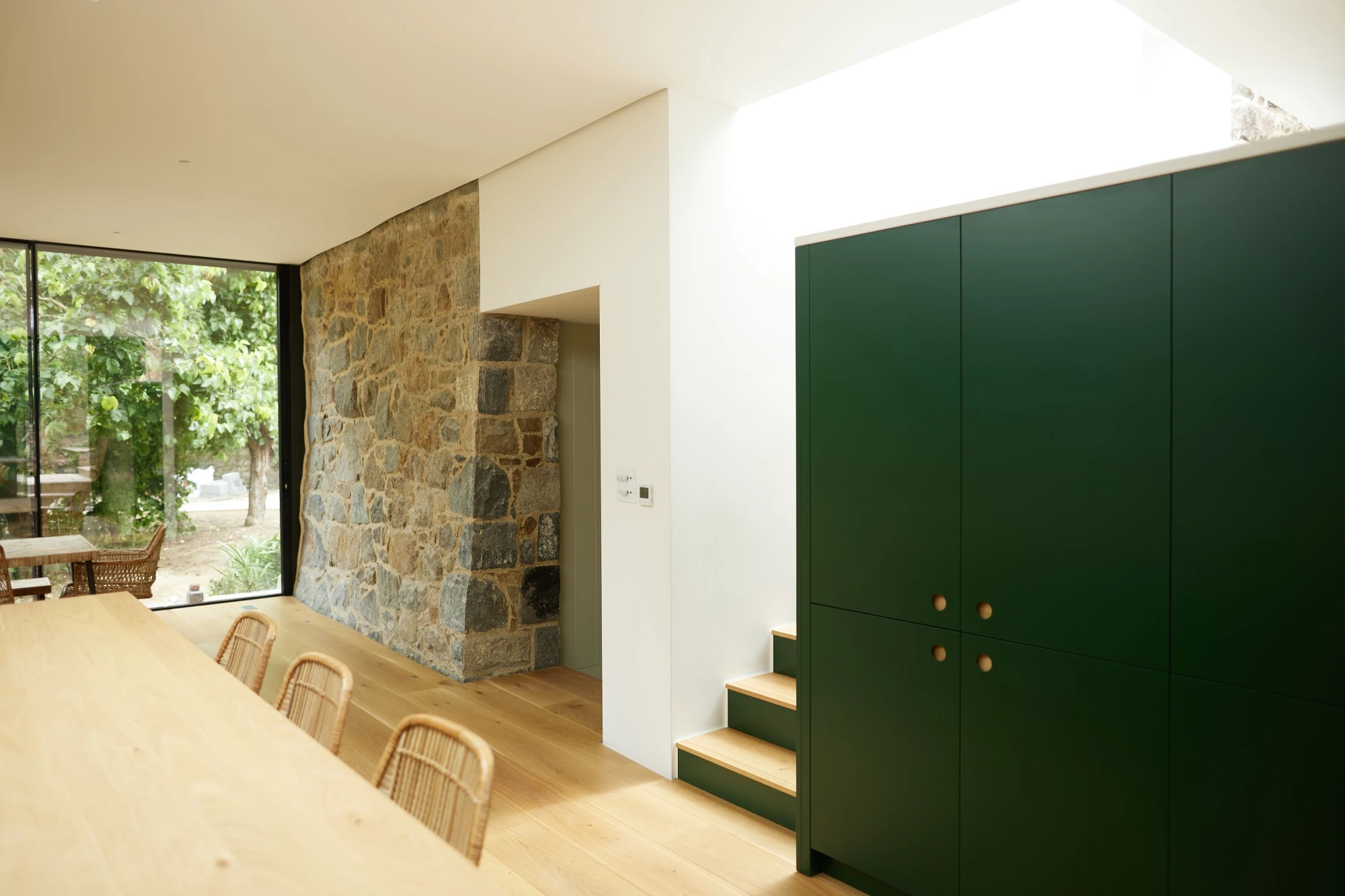 A modern kitchen featuring a minimalist design with light wooden flooring and green cabinetry. A stone wall adds a rustic touch. Large windows on the left side allow natural light to stream in, illuminating the space. Several wicker chairs are placed near a dining table.