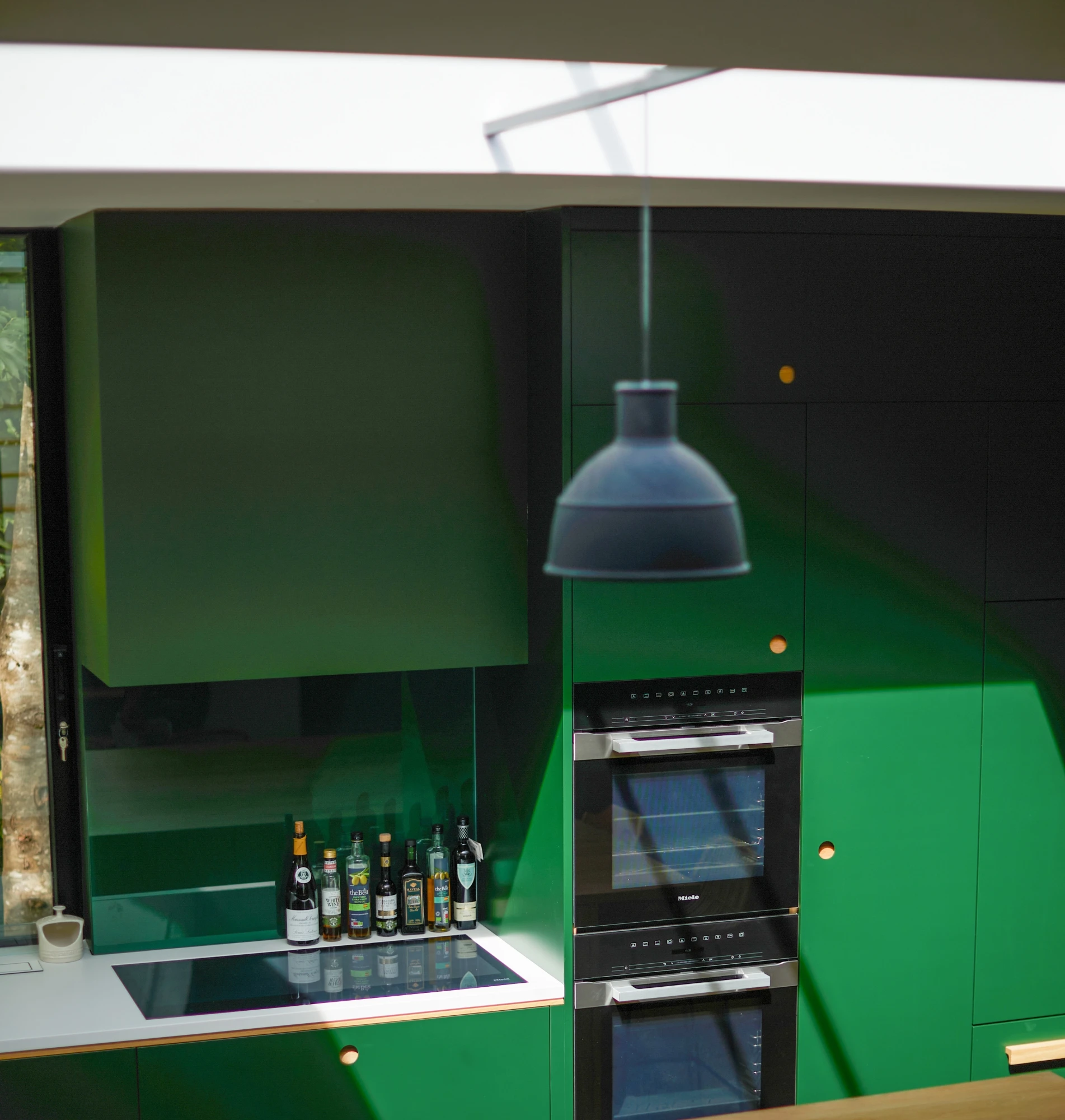 A modern kitchen with dark green cabinets and a built-in stove and oven. Bottles are arranged above the counter, beneath a black hanging light. Sunlight filters through a skylight, casting shadows on the cabinets.