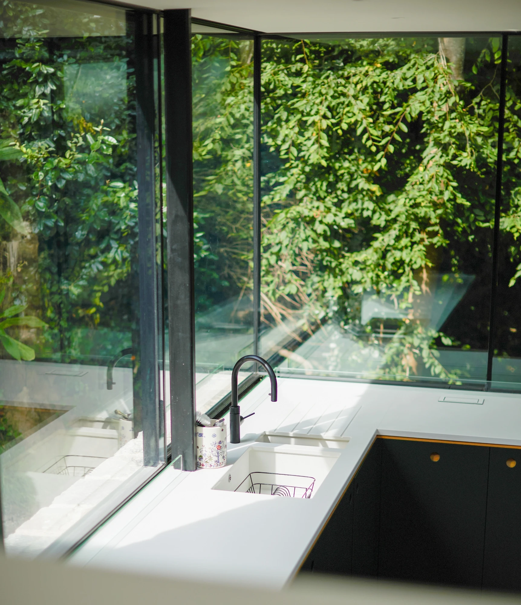 A modern kitchen features large floor-to-ceiling windows overlooking lush greenery. A black faucet and sink are installed on the white countertop, with a small container placed nearby