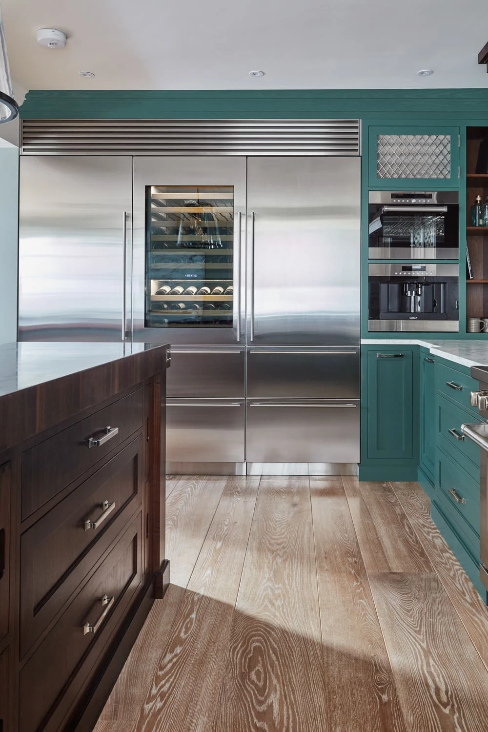 Modern kitchen featuring a large stainless steel refrigerator with a glass panel showcasing wine bottles. The cabinetry is green, and there's a built-in coffee machine on the right. Wooden flooring and a dark wooden island are also visible