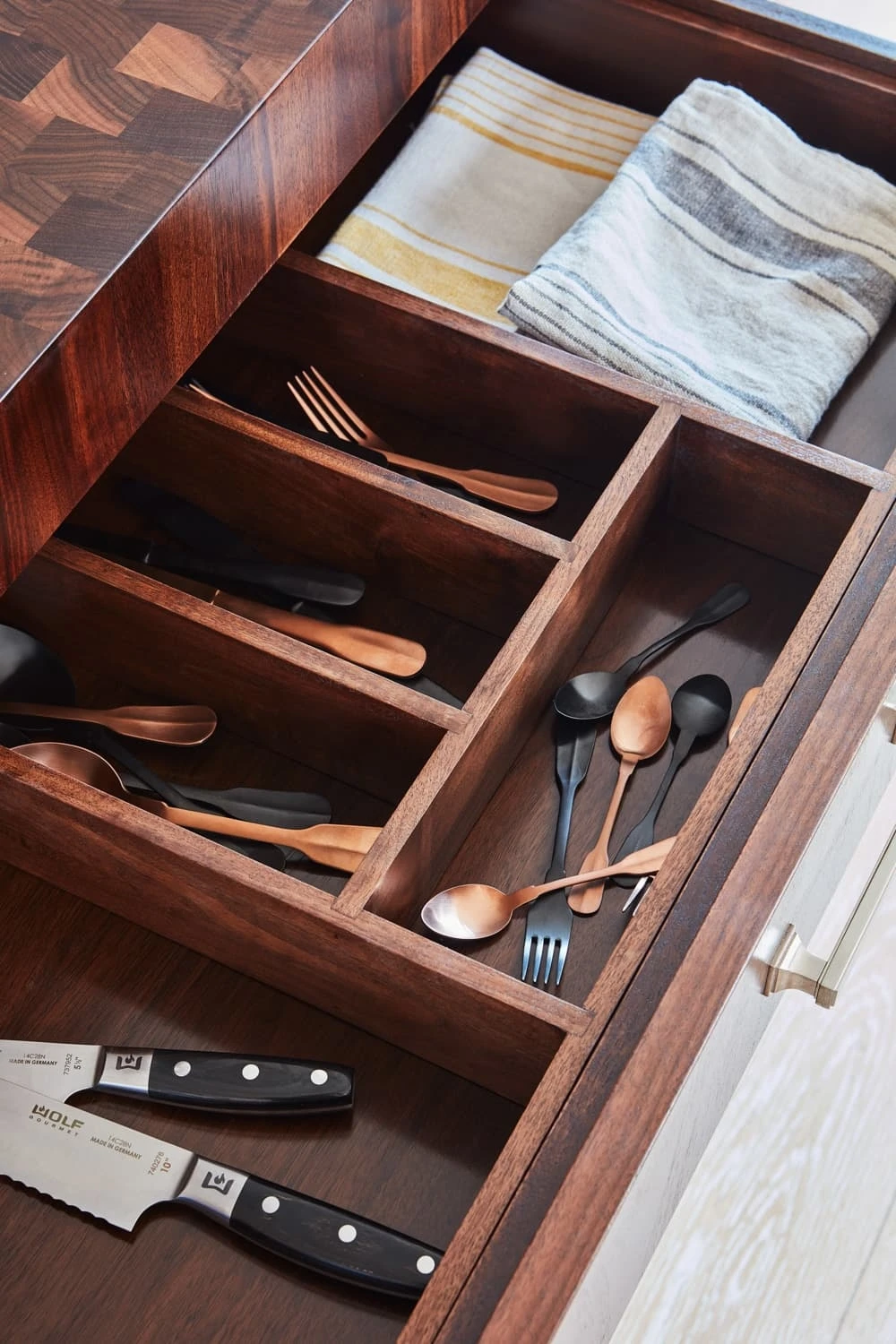 Open kitchen drawer with wooden dividers containing neatly arranged cutlery, including black and copper utensils. Two kitchen knives are in a separate section. Folded striped kitchen towels are visible in the top compartment