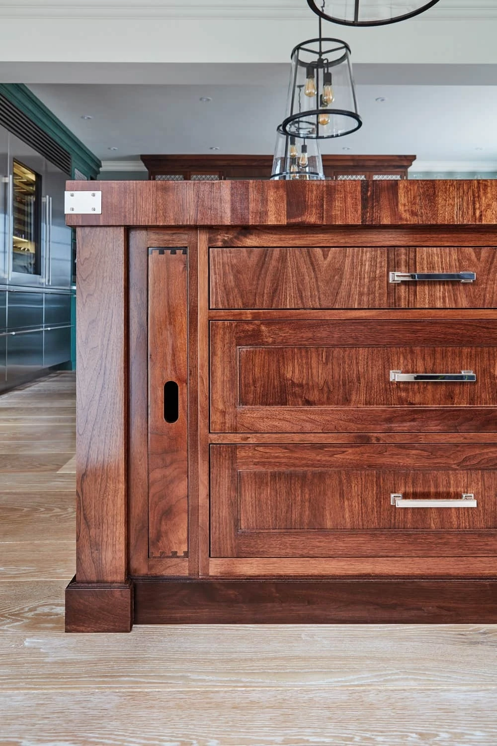 Close-up of a wooden kitchen island featuring rich brown tones and sleek metal handles. The design showcases a blend of rustic and modern elements, with three drawers and a side panel, set against a background of light wood flooring and hanging lights.