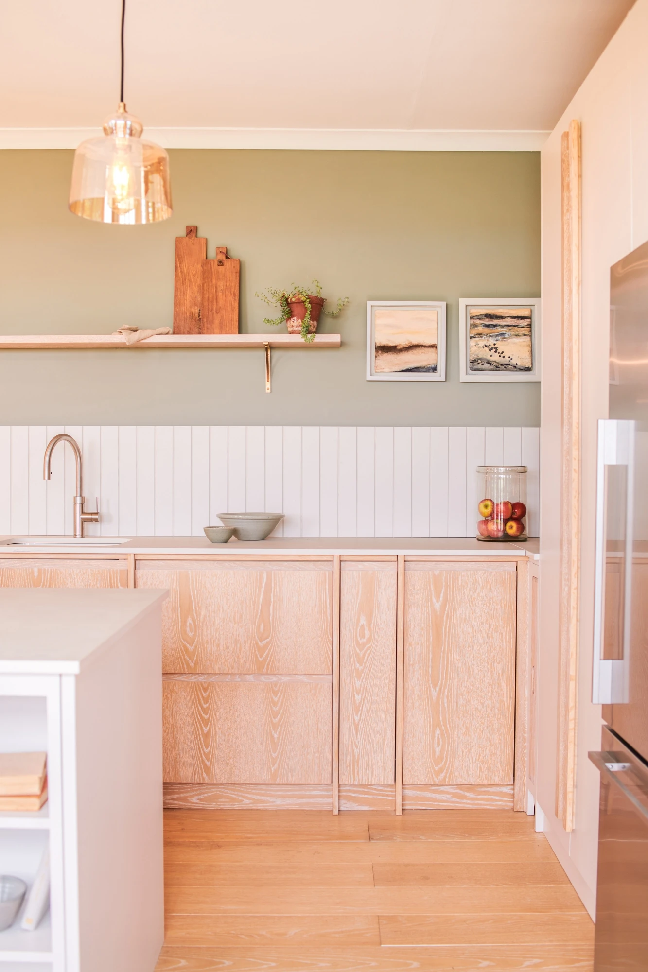 A modern kitchen features light wood cabinets with a natural grain, a white countertop, and a backsplash with vertical white tiles. The wall above is painted olive green, and a wooden shelf displays decorative items including a cutting board and vases. The floor is also light wood.