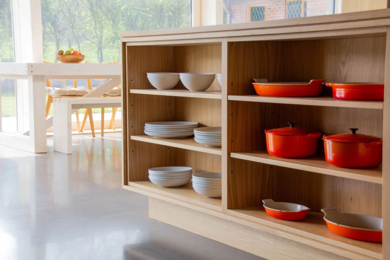 A wooden kitchen shelf displaying orange pots and white bowls and plates. Sunlight streams in, highlighting the minimalist design. A white dining table with a fruit bowl is visible in the background.