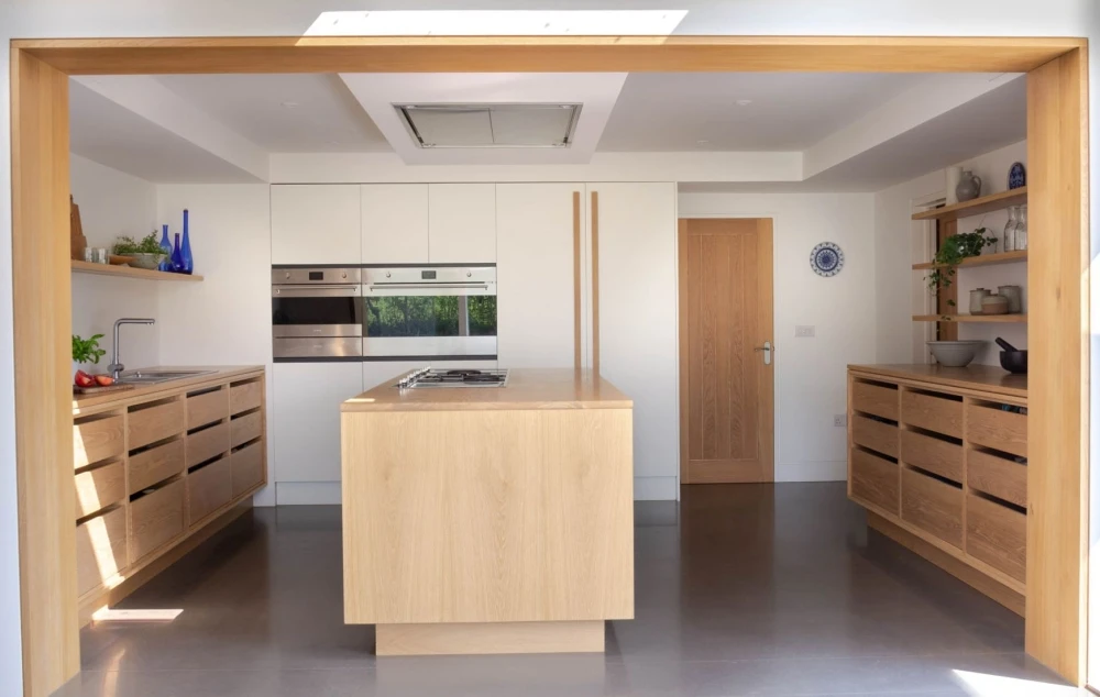 A modern kitchen with light wood cabinets and drawers, an island counter in the center, and stainless steel appliances. The kitchen has open shelving with various items and a door in the background. A skylight above lets in natural light.