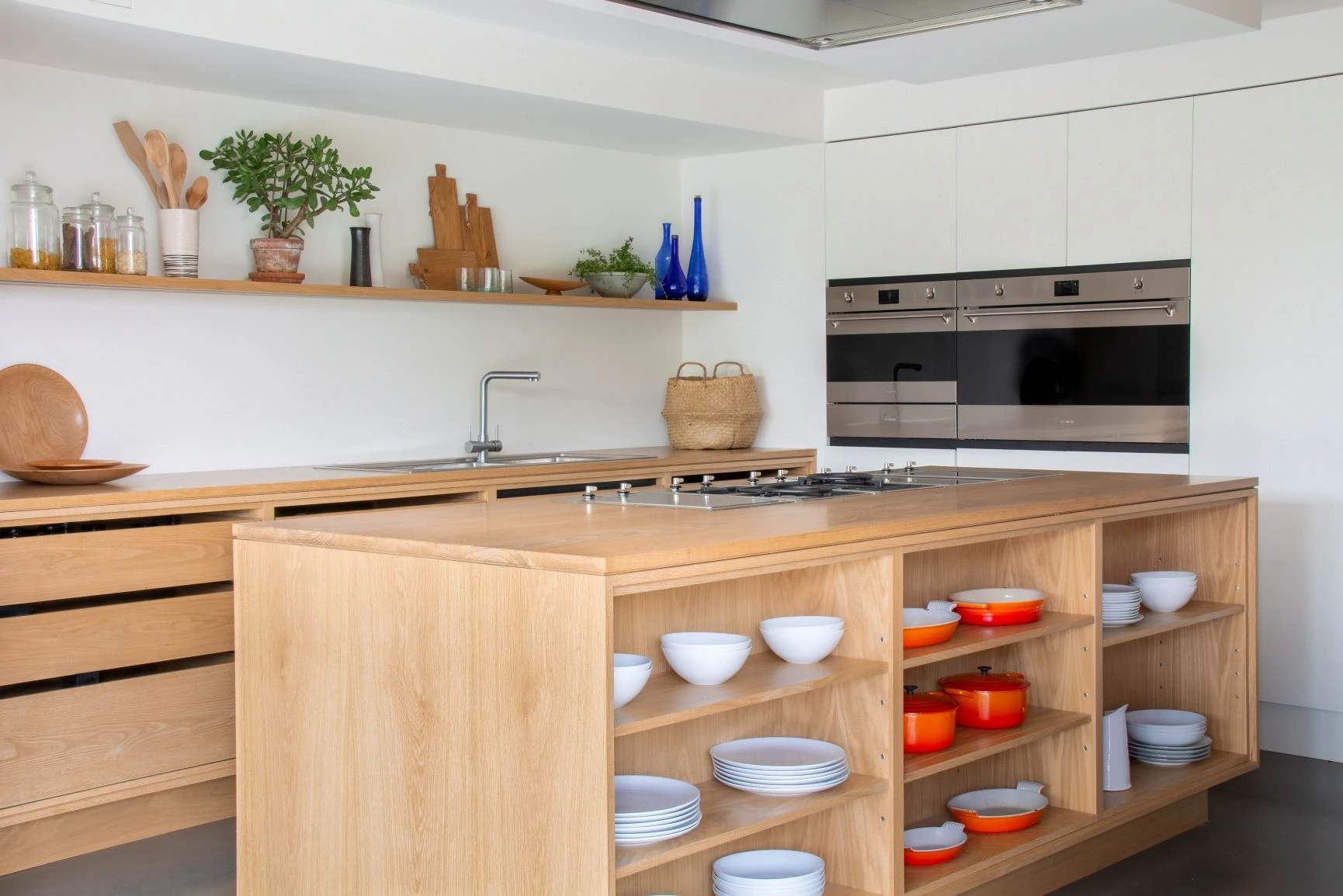 Modern kitchen with light wood cabinets and a spacious island. The island features open shelves with white and orange bowls. The countertop has a built-in stove. The back wall has a shelf with plants and blue bottles. Double ovens are integrated into the cabinets.