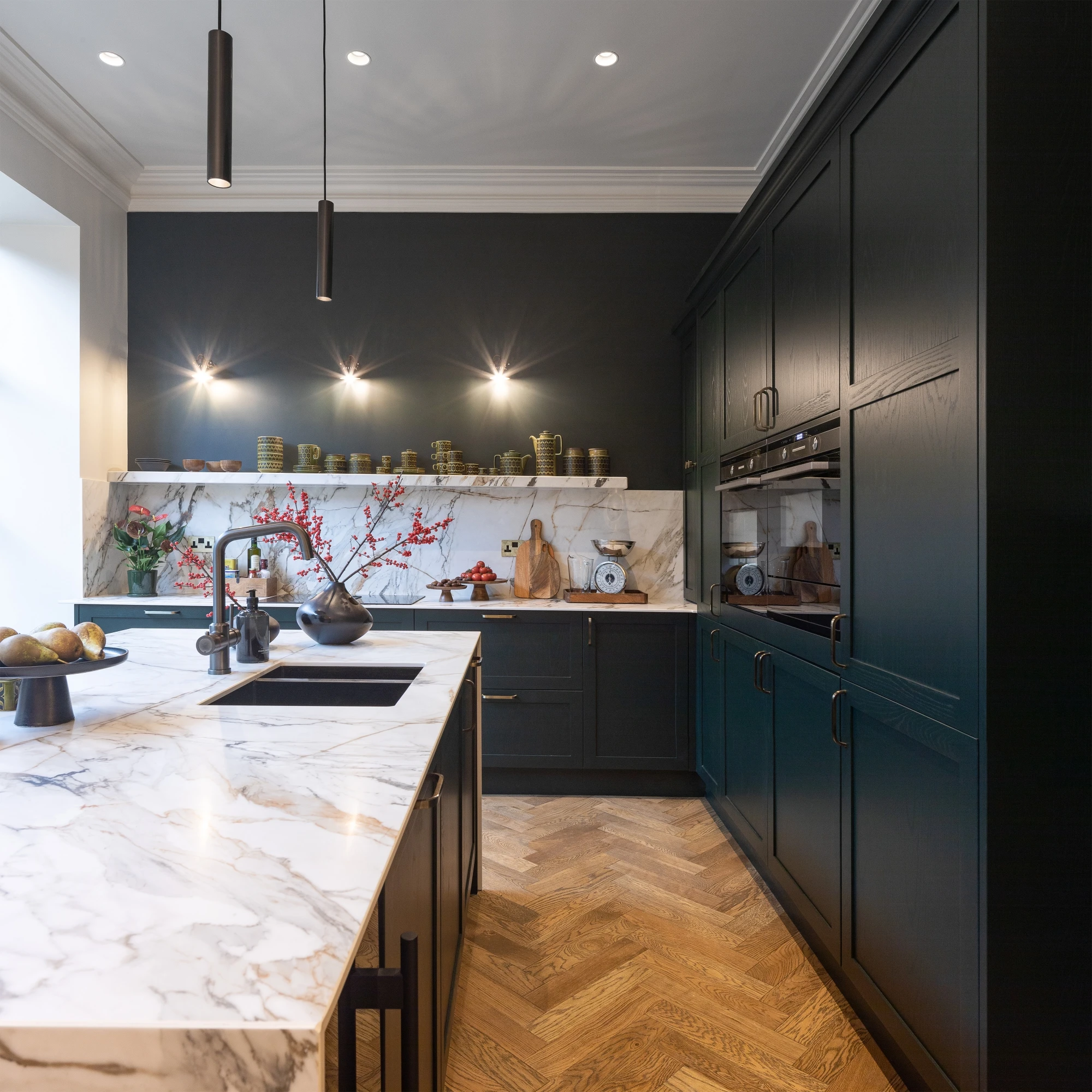 Modern kitchen with dark cabinetry, marble countertops, herringbone wooden floor, and elegant lighting