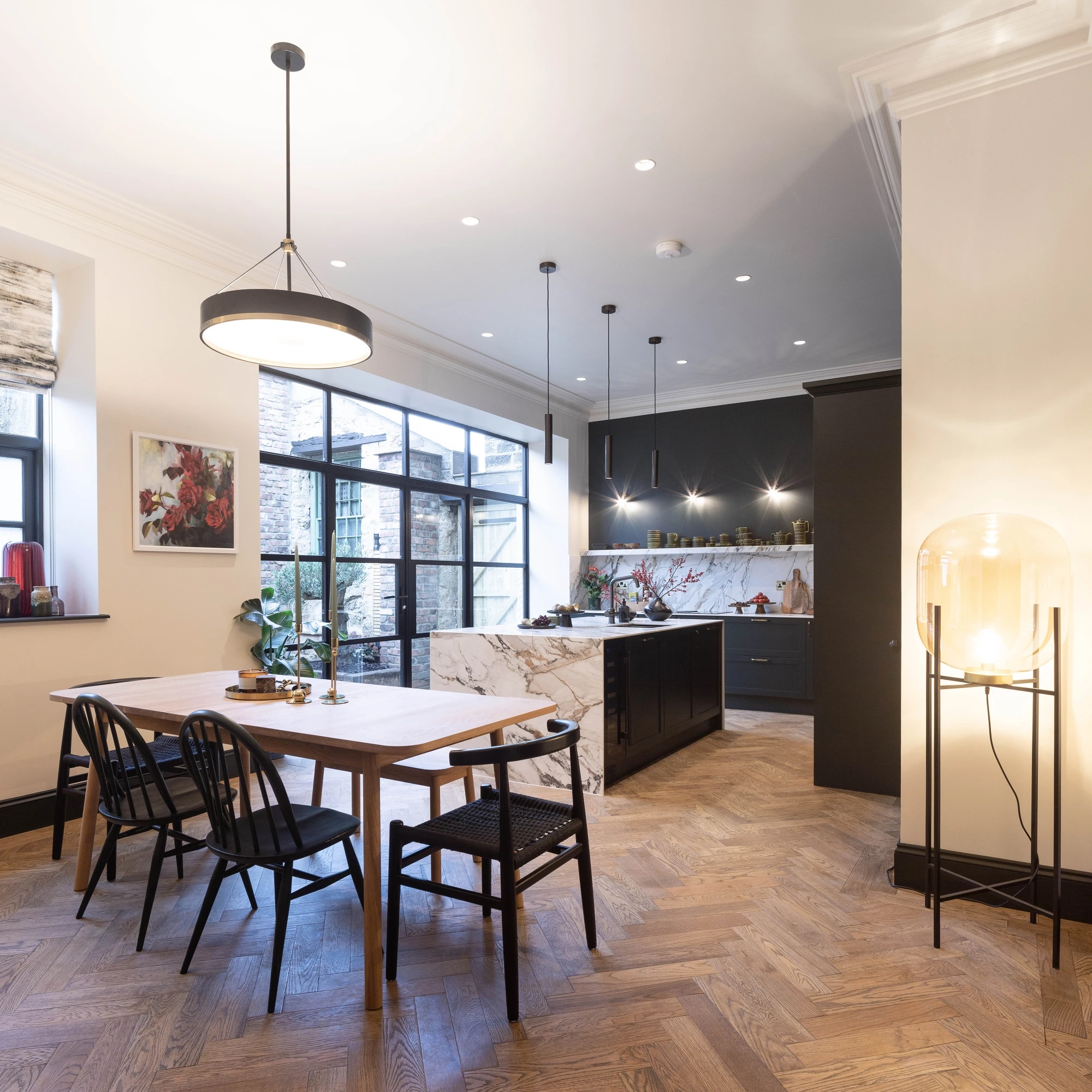 Open-plan kitchen and dining area with marble island, dark cabinetry, large windows, wooden herringbone flooring, and modern lighting