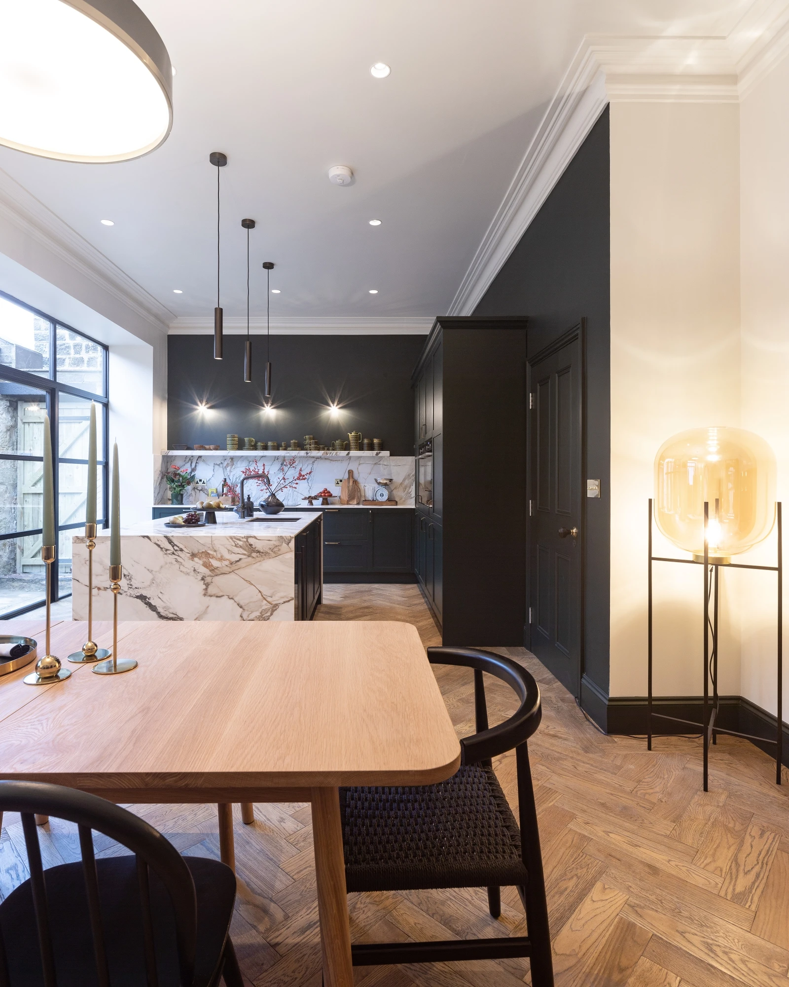 A modern kitchen and dining area with black cabinets, a marble island, and a wooden dining table with black chairs. The room features herringbone wood flooring, large windows letting in natural light, hanging pendant lights, and a tall floor lamp with a soft glow