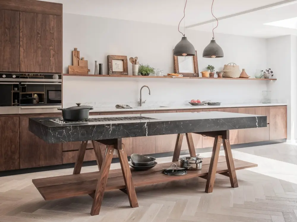 A modern kitchen featuring a large marble island with a black cooktop and wooden lower shelf. The background has wooden cabinets, a marble backsplash, a stainless steel oven, and minimalist decor including framed pictures, plants, and two pendant lights.