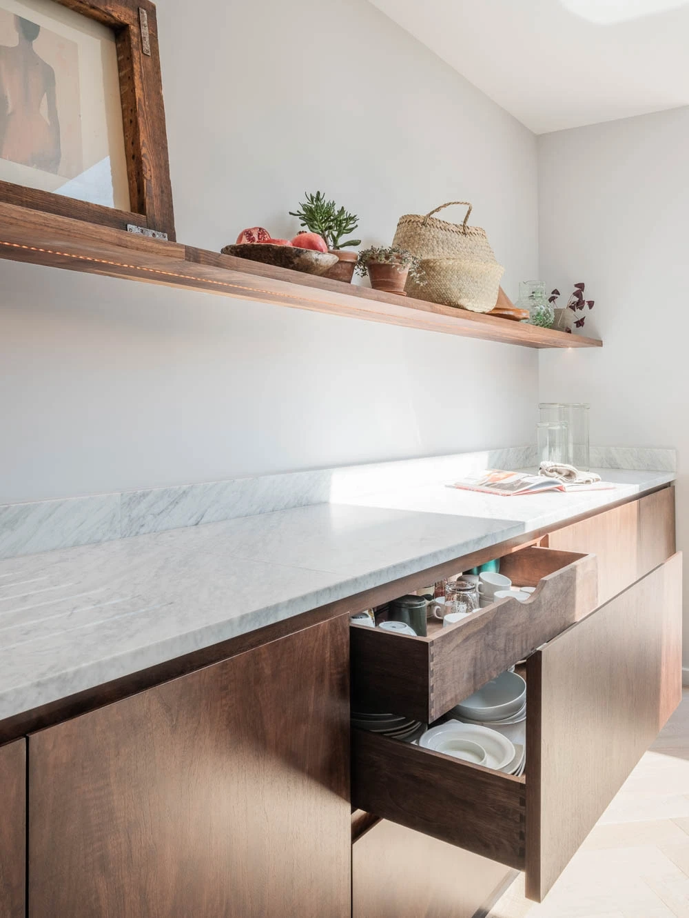 A modern kitchen with a marble countertop, open drawers displaying dishware, and a wooden shelf above. The shelf holds decor items like potted plants and a woven basket. Sunlight highlights the clean, minimalist design