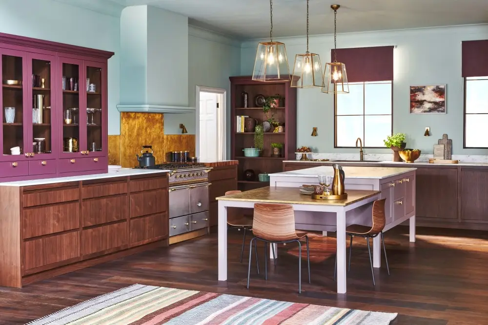 A spacious kitchen with light wood flooring and a dining area featuring wooden chairs and a white table. The kitchen has two-tone cabinets, one island with shelves and a wine cooler, and another with open shelving. Large windows brighten the space.