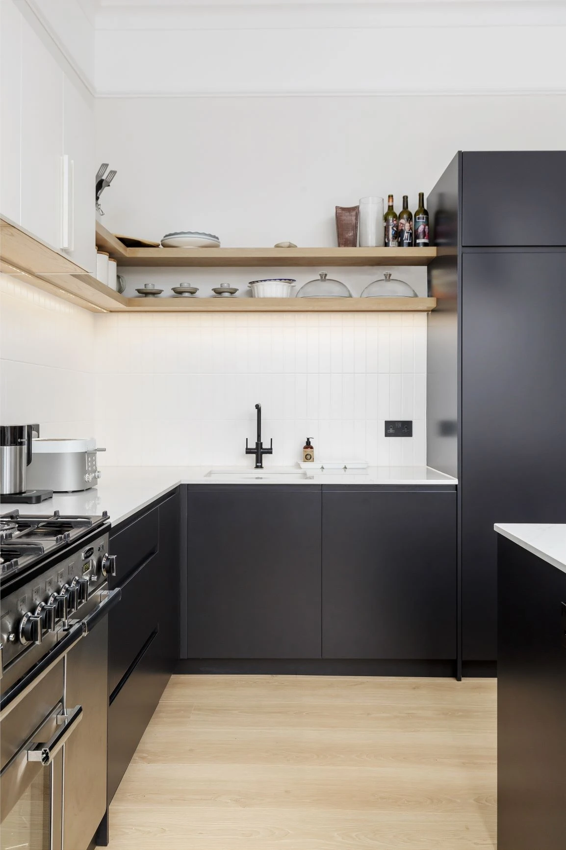 A modern kitchen with black lower cabinets and white upper cabinets. There are open wooden shelves holding various kitchen items and bottles. The countertops are white, and there is a black sink faucet. The floor is light wood and there is a stainless steel stove.