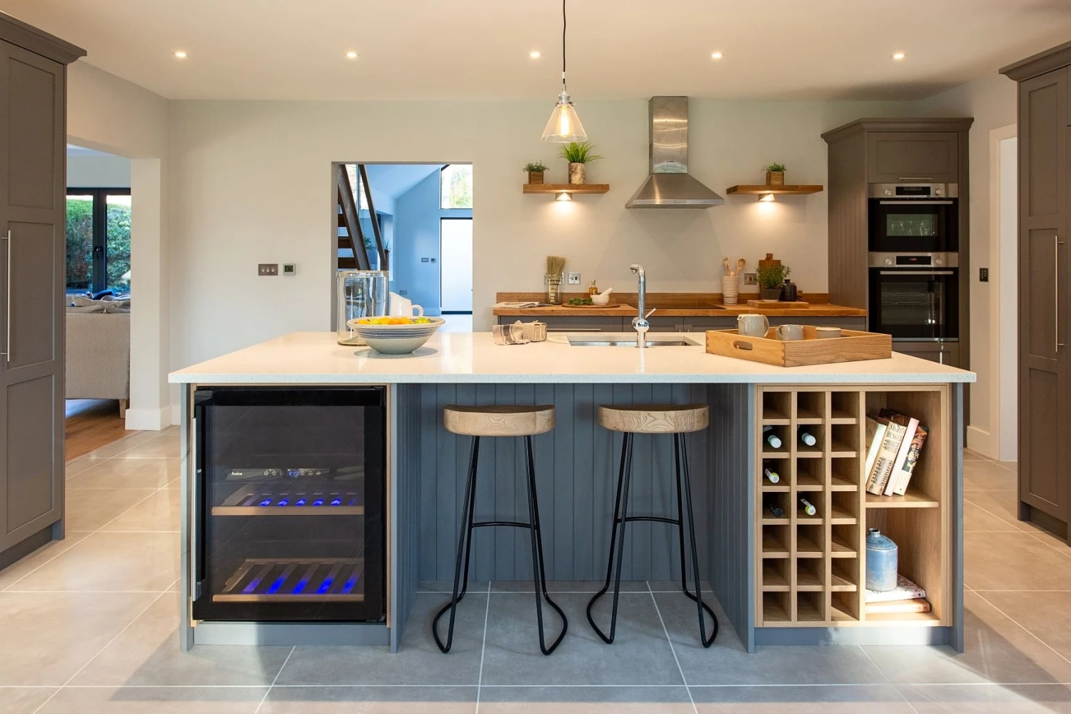 Modern kitchen with a large island featuring two wooden bar stools and built-in wine storage. The island has a countertop sink. There's a wine cooler with a glass door. Stainless steel appliances are in the background, and the room is well-lit.