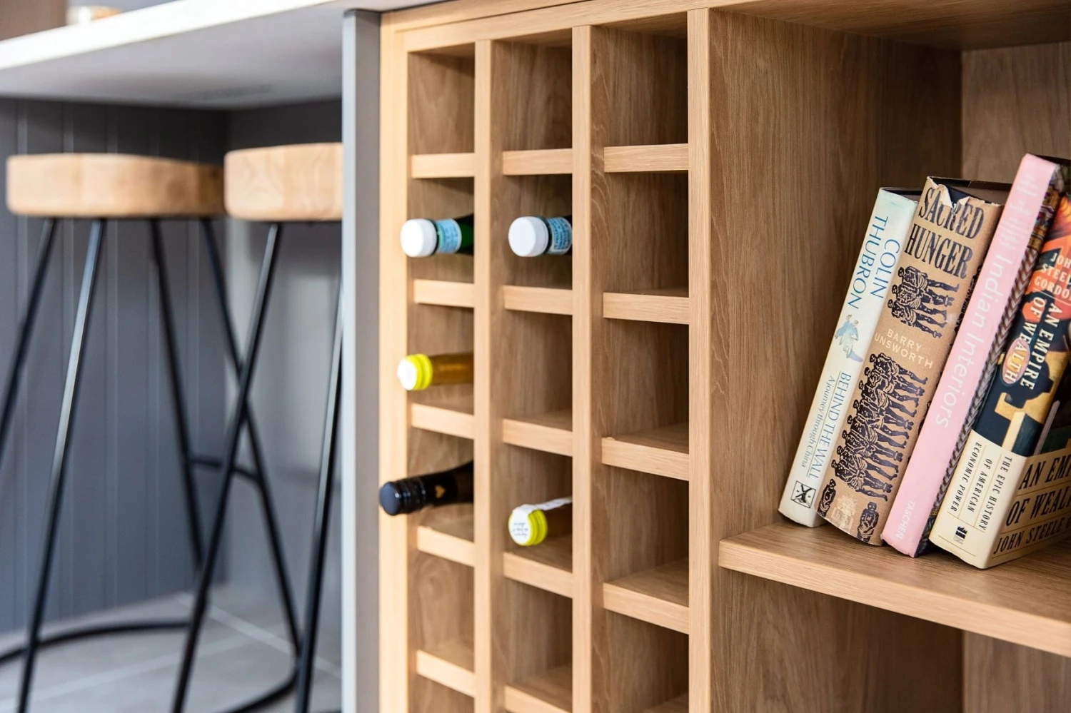 A wooden wine rack with several wine bottles and a few books on the right. Two wooden bar stools with black metal legs are in the background near a kitchen counter.