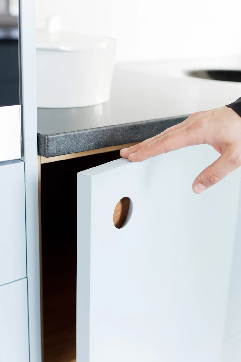 A person opens a white cabinet door with a Ladbroke handle below a dark countertop in a modern kitchen.