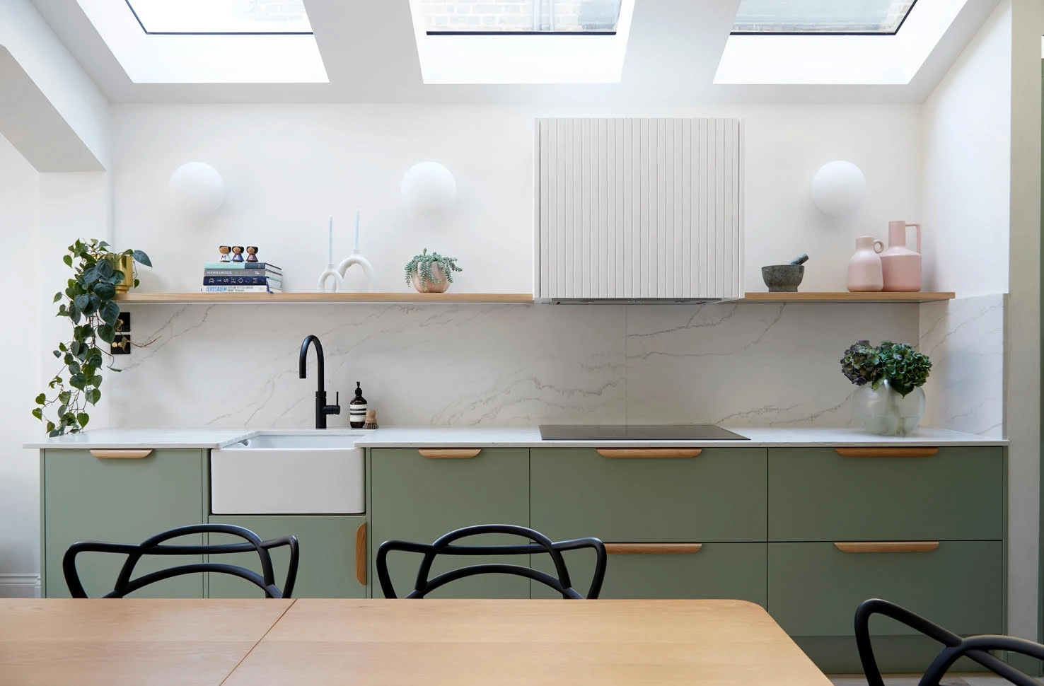 A modern kitchen featuring green lower cabinets with wooden handles, a white farmhouse sink, black faucet, a smooth white countertop, and a minimalist backsplash. A floating shelf holds decor items and plants. Skylights provide natural light above the counter