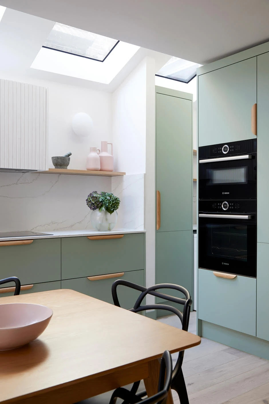 A modern kitchen with light green cabinets, marble countertops, and black appliances. There's a wooden table with black chairs in the foreground, a pink bowl on the table, and a shelf adorned with potted plants and decorative items. Skylights are overhead.