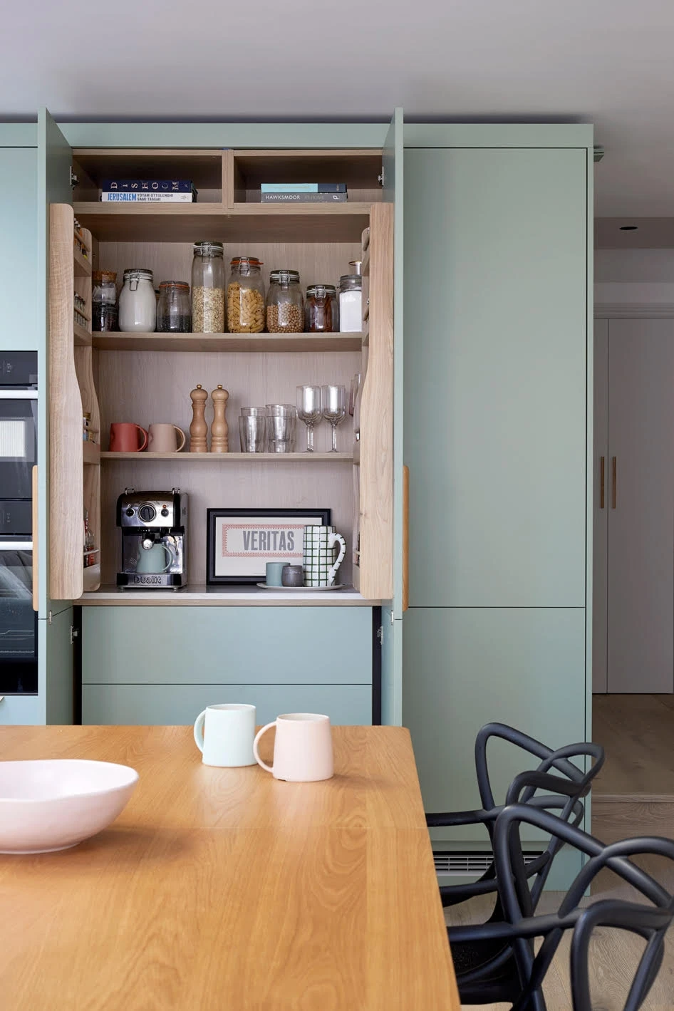 A modern kitchen featuring a light mint-green cabinet with wooden handles, a wooden table with a light pink bowl and two pastel-colored mugs (one mint green and one light pink). In the background, there are black chairs with a distinctive design.