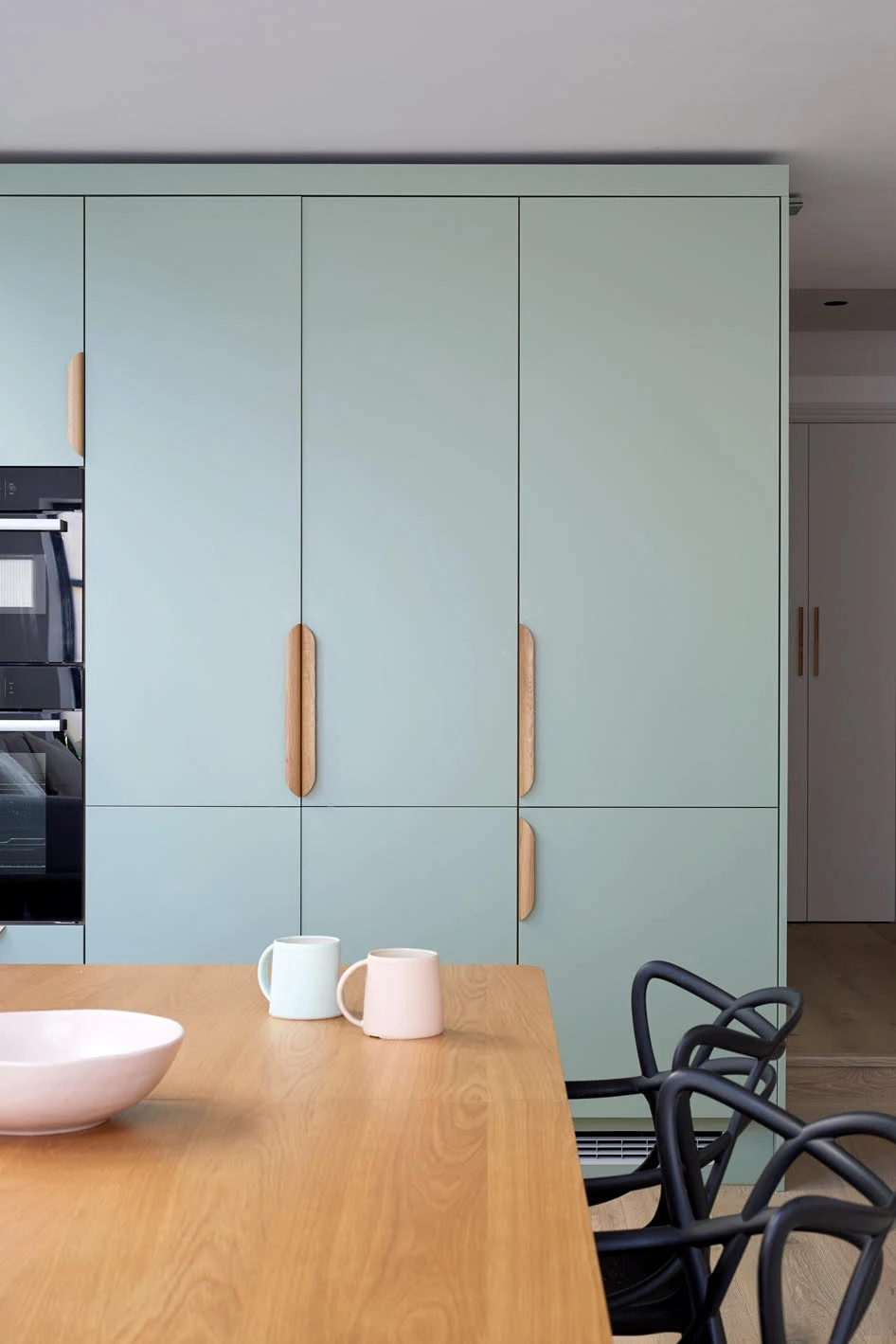 A modern kitchen featuring a light mint-green cabinet with wooden handles, a wooden table with a light pink bowl and two pastel-colored mugs (one mint green and one light pink). In the background, there are black chairs with a distinctive design.