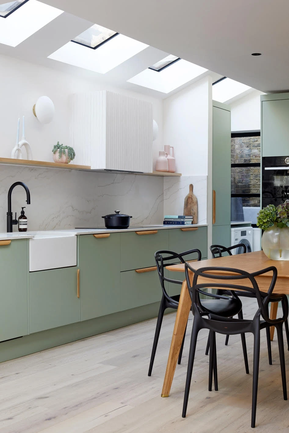 Modern kitchen with light green cabinets, white countertops, and a farmhouse sink. Black faucet and kitchenware are on the counters. Light wood dining table with black chairs is in the foreground. Skylights and light wood flooring brighten the space.