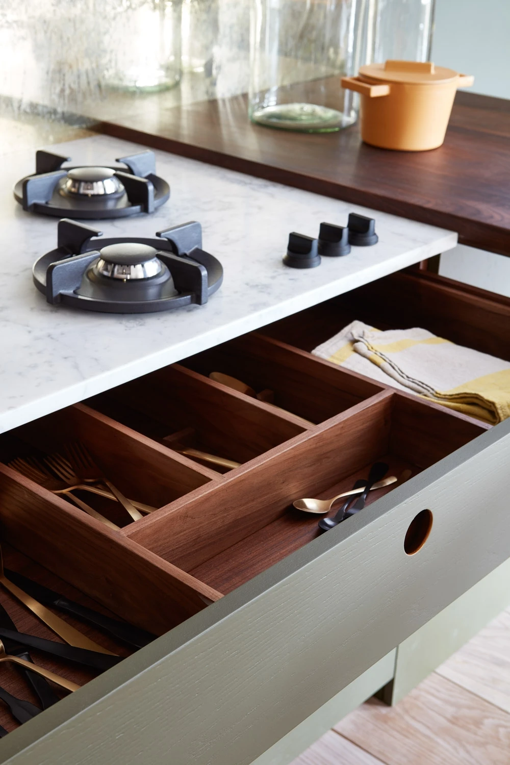 A kitchen countertop with a two-burner gas stove and open wooden cutlery drawer underneath. The drawer holds utensils and a folded yellow cloth, with a marble surface and jars in the background.