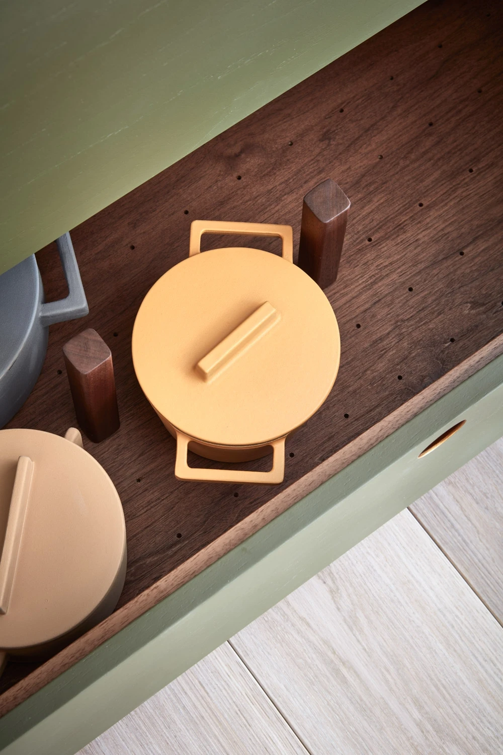 A neatly organized shelf features two cast iron pots with lids, one light beige and the other mustard yellow. The shelf has a wooden base with wooden dividers, placed in a green cabinet with a handle