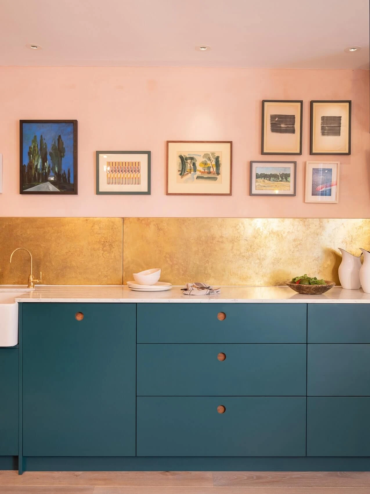 A modern kitchen with teal cabinets, gold backsplash, and a white countertop. The wall is decorated with various framed artworks against a light pink backdrop. There are a few dishes and greenery on the countertop.