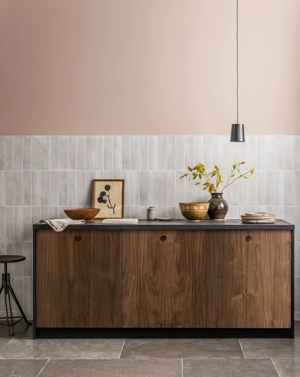 Modern kitchen featuring a wooden cabinet with a dark countertop. On the countertop, there are various ceramic bowls, plates, and a plant in a vase. A minimalist framed picture leans against a light-colored tile backsplash. A black pendant light hangs above.