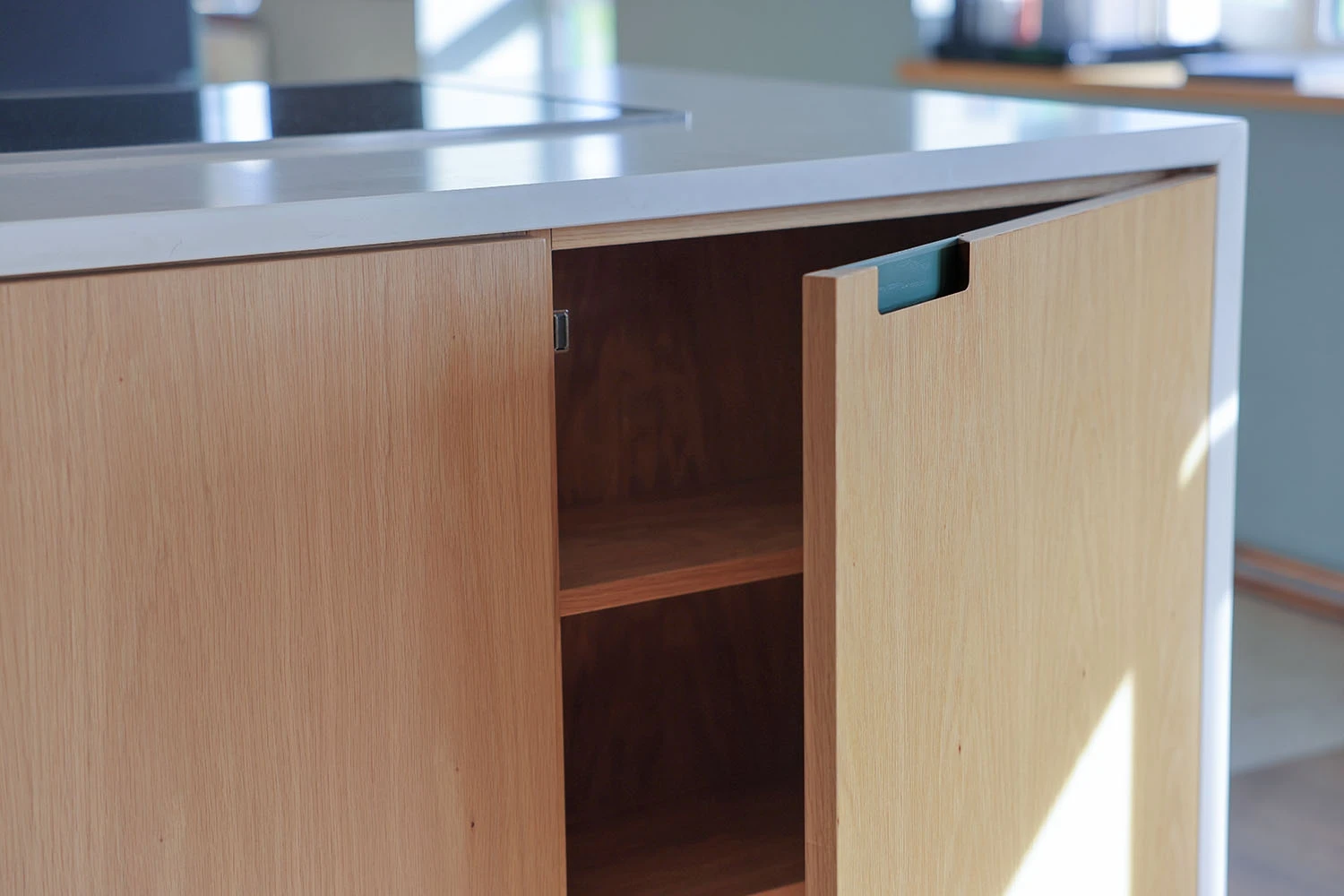 A modern kitchen cabinet with a wooden finish is shown with its door slightly ajar, revealing two empty shelves inside. The countertop above the cabinet has a sleek, minimalist design. Sunlight casts shadows on the surface.