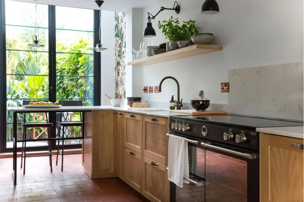 A modern kitchen features wooden cabinets with a black stovetop and oven. A white towel hangs on the oven handle. A large window offers a view of lush greenery. Open shelves hold potted plants and kitchenware. The floor is tiled with reddish-brown tiles