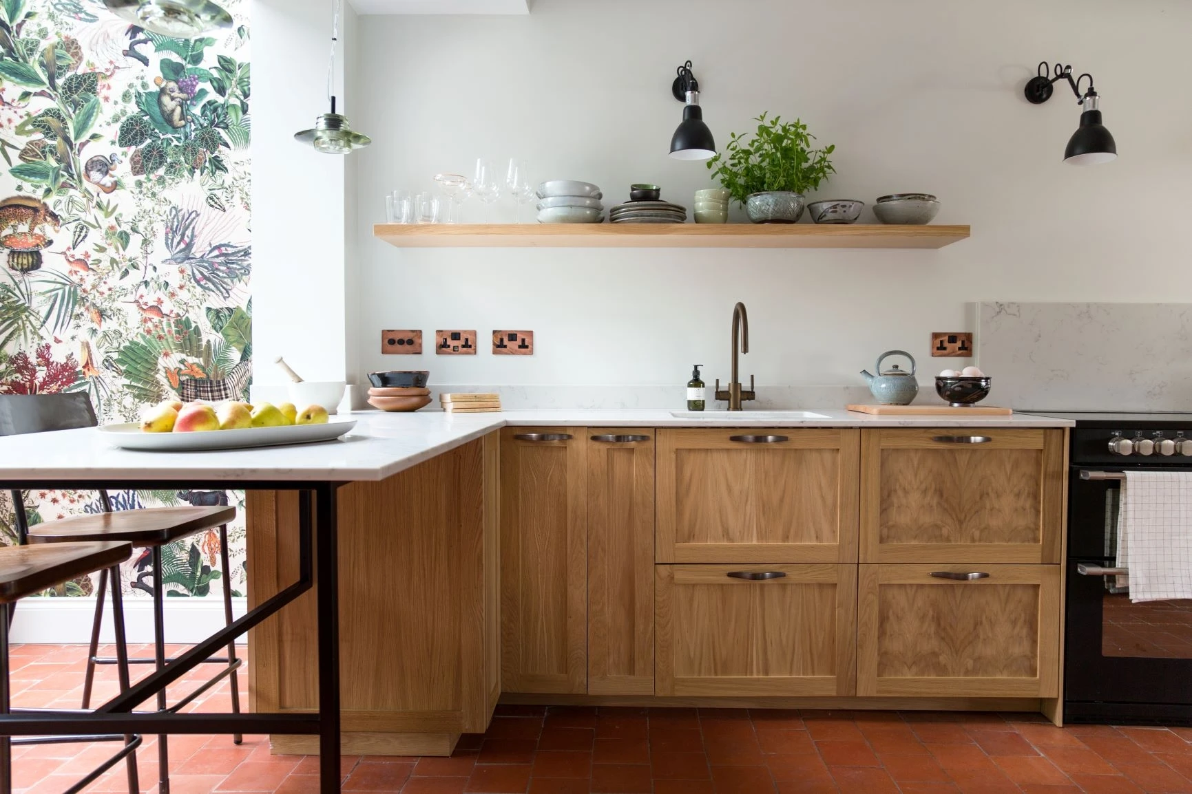 A modern kitchen features light wood cabinets, a white countertop, and black fixtures. Open shelving holds dishes and plants. A bowl of fruit sits on a black metal and wood island with stools. A vibrant floral wallpaper decorates one wall, adding color to the space.