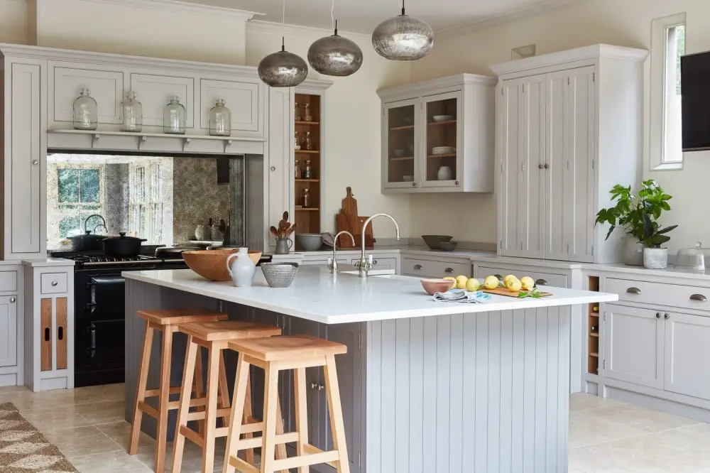 A spacious kitchen with a large island featuring a white countertop and wooden stools. Grey cabinets surround the space, and pendant lights hang above. A plant and fruit bowl decorate the counter, creating a modern and inviting atmosphere.