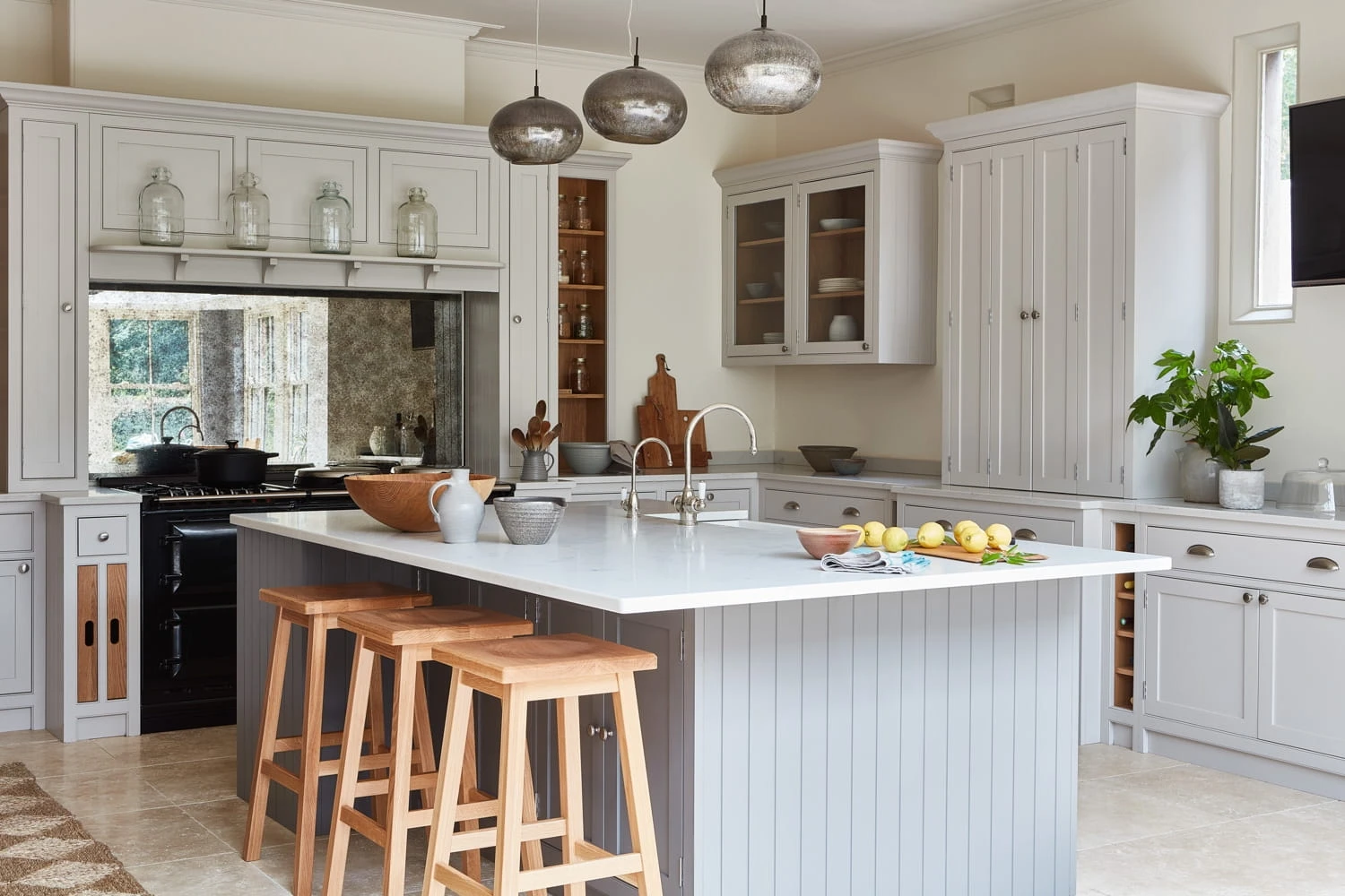 A spacious kitchen with a large island featuring a white countertop and wooden stools. Grey cabinets surround the space, and pendant lights hang above. A plant and fruit bowl decorate the counter, creating a modern and inviting atmosphere.