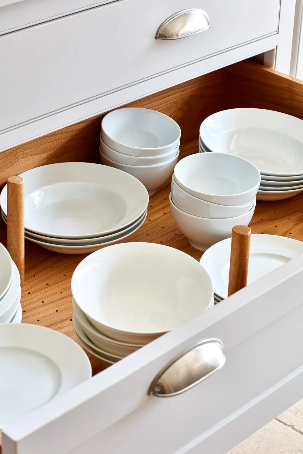 Open kitchen drawer with neatly organized white ceramic bowls and plates. Plates are stacked horizontally, while bowls are in groups, showcasing a tidy arrangement. The drawer has a wooden interior with adjustable pegs for customization.