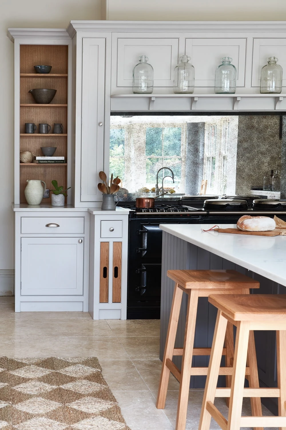 A modern kitchen with white cabinets, open shelving displaying dishes, and a black stove. A large marble countertop with wooden bar stools is in the foreground. Glass jars adorn the cabinet top, and a woven rug lies on the tile floor.