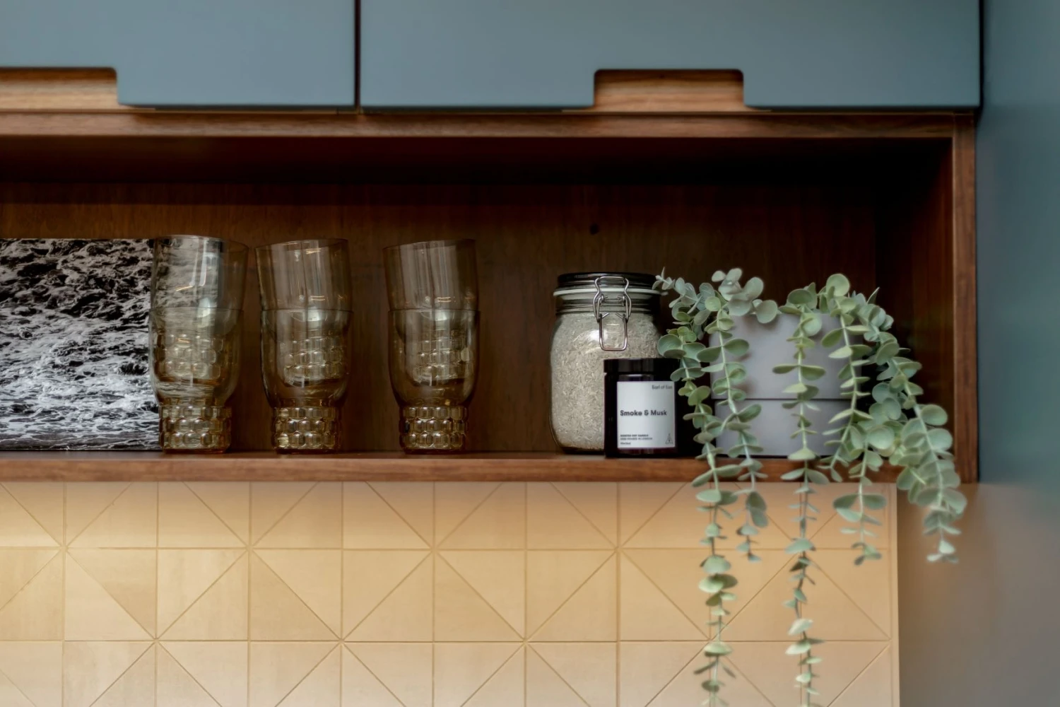 A wooden kitchen shelf displays translucent glasses and a jar labeled 