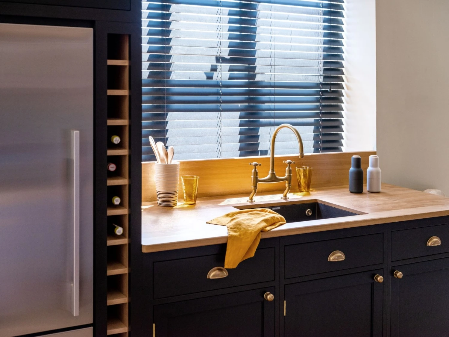 A modern kitchen with black cabinets, a wooden countertop, and a brass faucet over a sink. There's a silver fridge on the left, wine bottles in the rack, and items like utensils and bottles near the window with blinds.