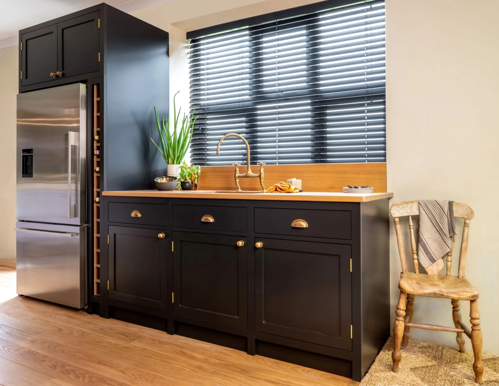 A modern kitchen with black cabinets adorned with gold handles, a stainless steel refrigerator, a wooden chair draped with a cloth, and a potted plant by the window. Wooden floors complement the sleek design. The window features black horizontal blinds.