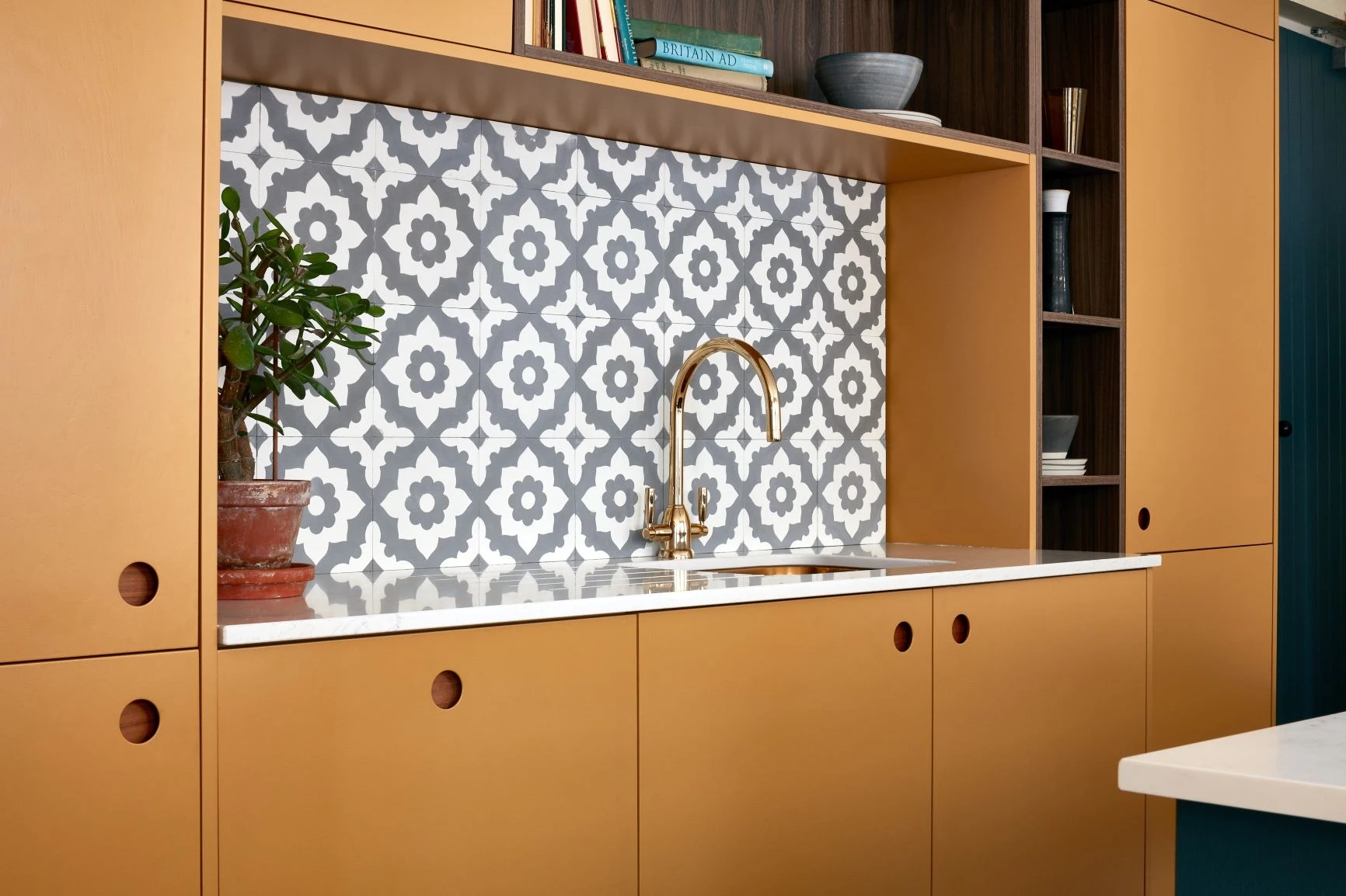 A modern kitchen features mustard-yellow cabinets with circular handles, a white countertop, and a decorative gray and white geometric tile backsplash. A potted plant sits beside a sleek brass faucet under open shelving with books and bowls