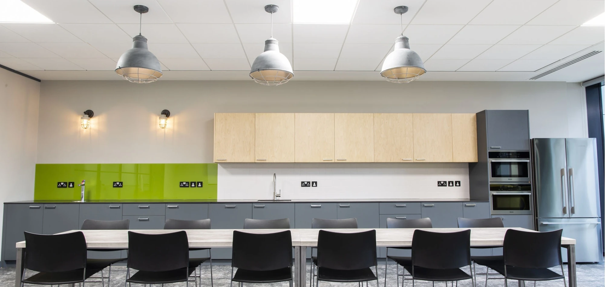 A modern kitchen with green backsplash, wooden cabinets, and stainless steel appliances. There are three industrial-style pendant lights above a long table with black chairs. The room has a minimalist and contemporary design.