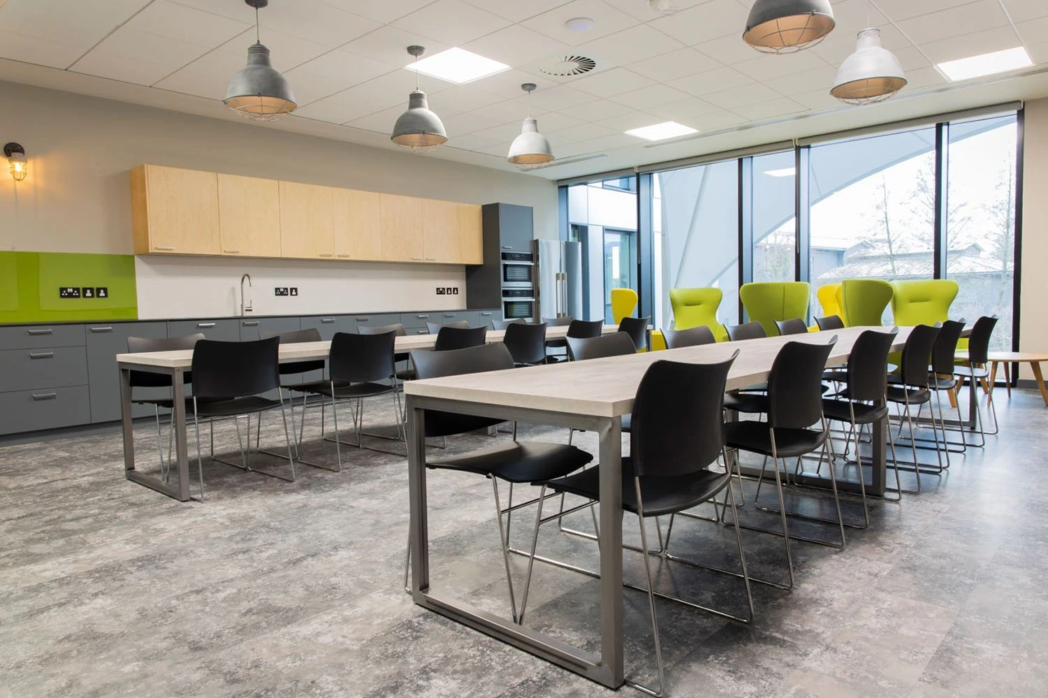 A spacious, modern cafeteria with large windows and natural light. It features long tables with black chairs, green accent walls, and pendant lights. There are lime green chairs near the window, creating a bright, inviting atmosphere.