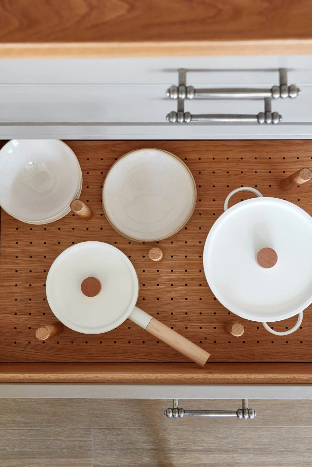 Open wooden kitchen drawer with pegboard base containing neatly arranged white ceramic bowls and cookware with wooden handles. Drawer features silver handles.