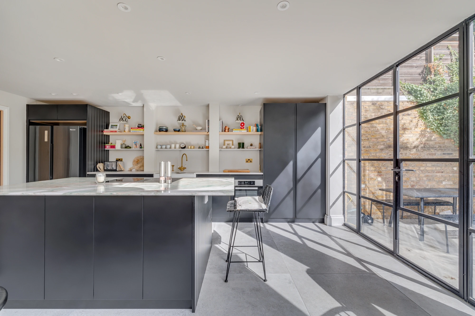 Modern kitchen with black cabinets and a large island with bar stools. Open shelving displays various kitchen items. Large industrial-style windows allow natural light to flood in, revealing an outdoor dining area with a brick wall and greenery