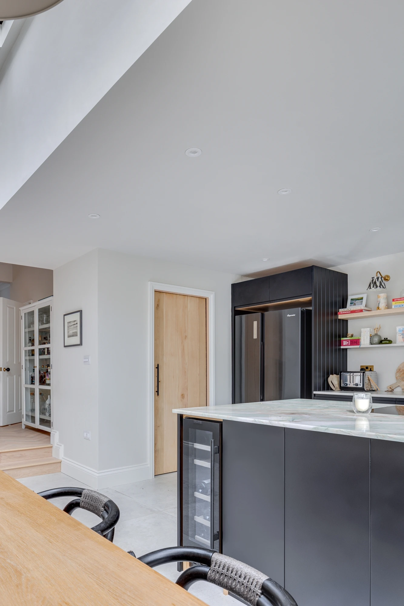 Modern kitchen interior with sleek black cabinets and a large refrigerator. A light wood door is seen in the background. Open shelving holds various kitchen items, and a dining table with chairs is visible in the foreground. The overall space is bright and airy.